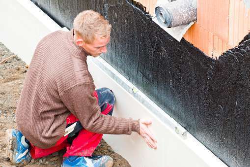 Construction worker installing styrofoam insulation board on a basement foundation and measuring with a spirit level. This image is part of a series.